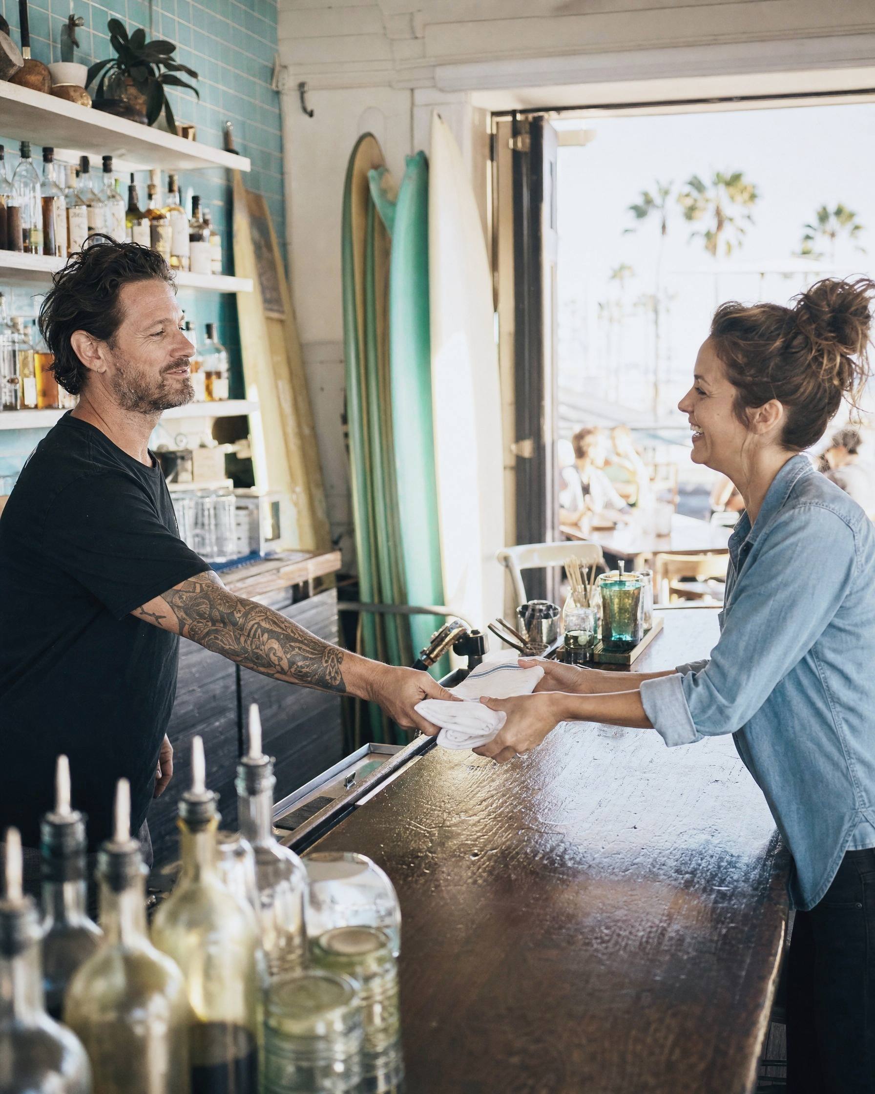 Behind the counter of a beach bar, the owner slides a folded bar towel to the woman taking over.