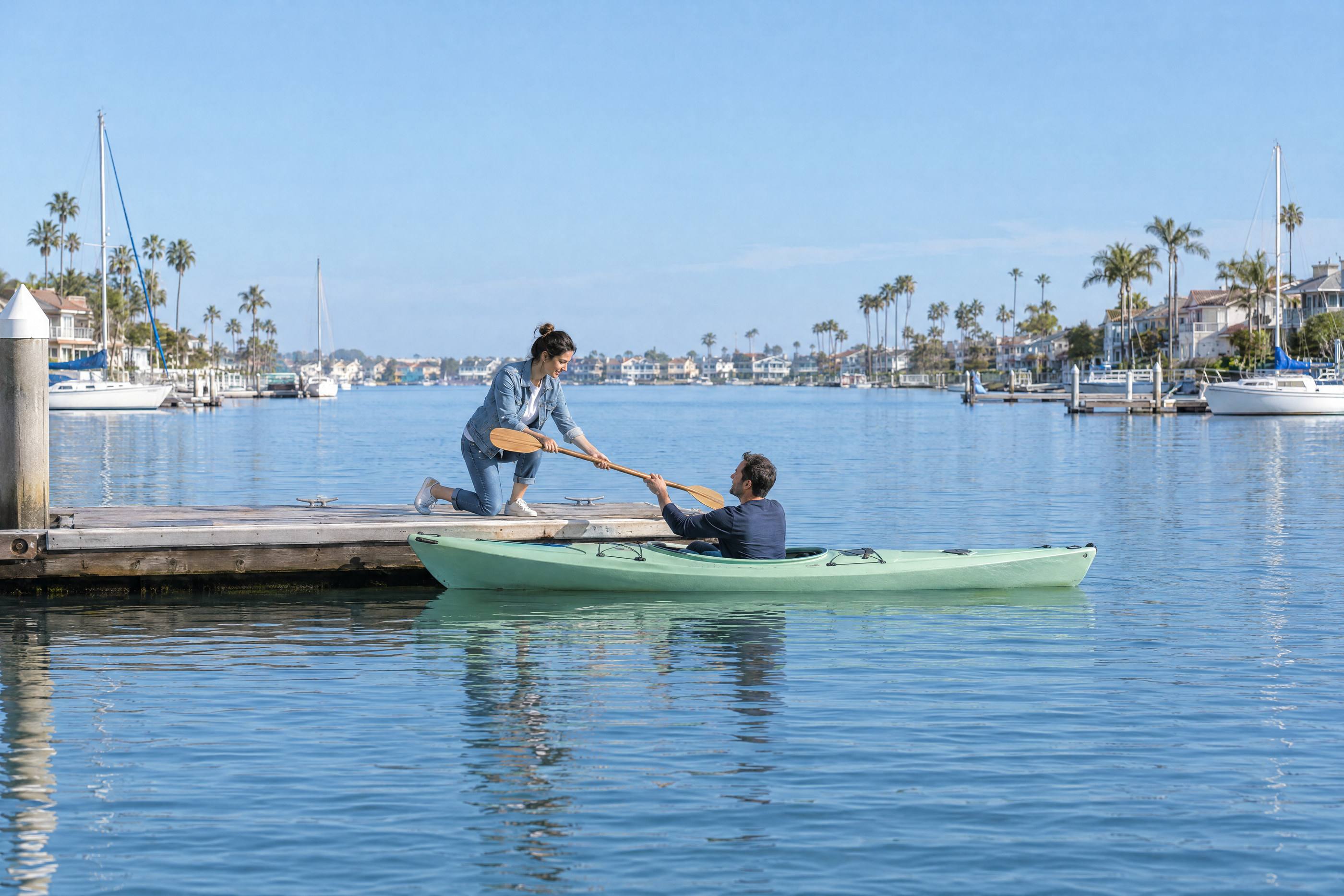 At the Huntington Harbour docks, a woman kneels on the dock and hands a wooden paddle to a man in a mint kayak.